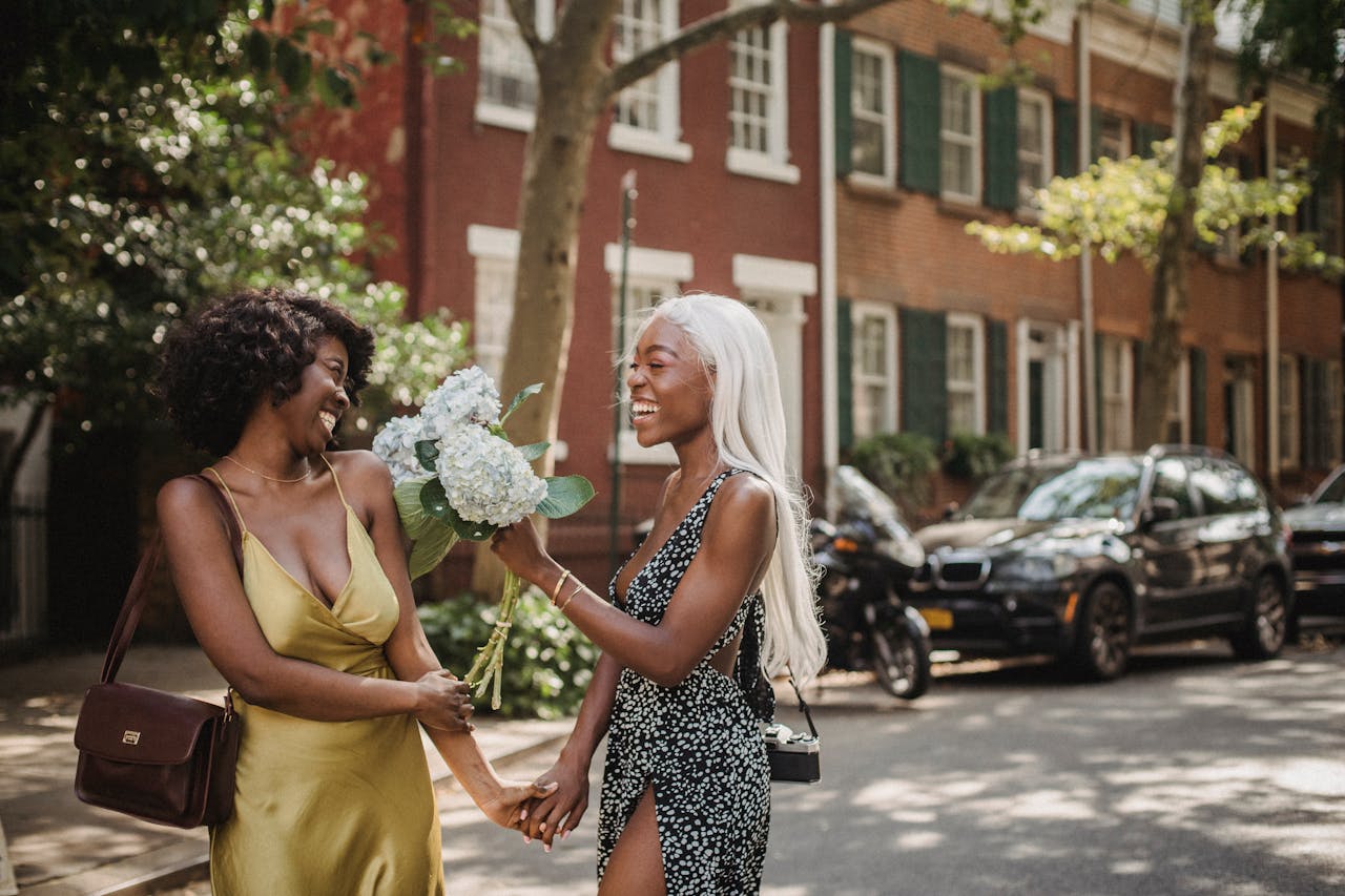 Two women enjoying a walk on a sunny street, holding hands and a bouquet of flowers, sharing smiles and happiness.